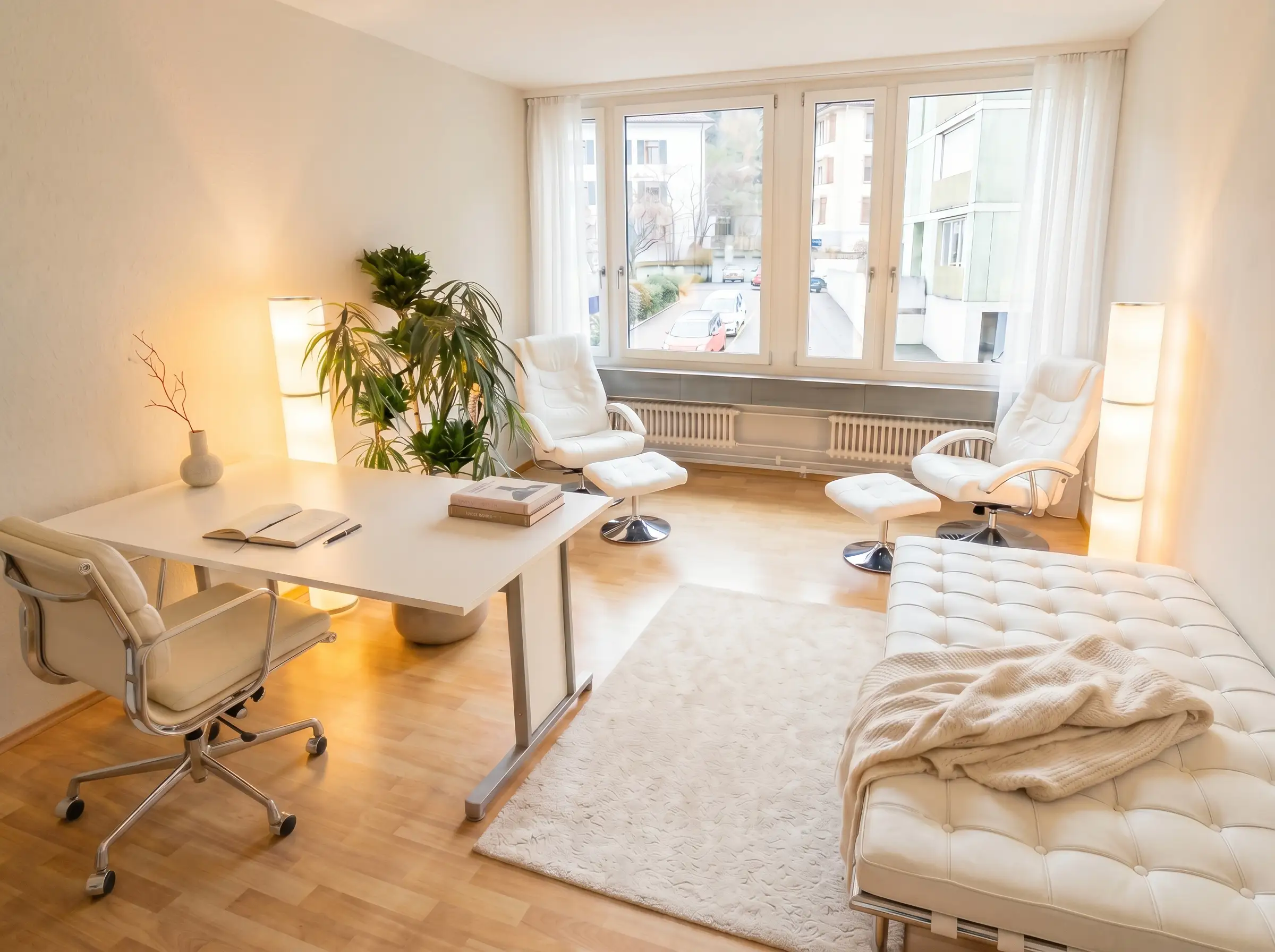 Der Praxisraum in Baden: Schreibtisch, zwei Sessel und die Liege am Fenster, warmes Licht